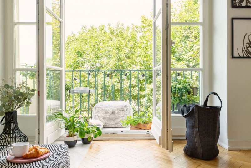 Raumheilung, offene Glastür von einem Wohnzimmer auf einem sonnigen Balkon mit grünen Pflanzen und bequemen Möbeln, großer Beutel aus groben und grauen Stoff