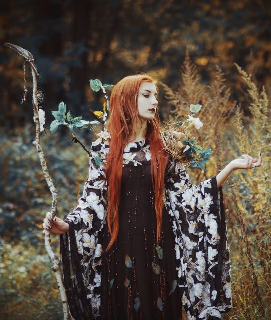 Energetisches Feng Shui, Völva, Stab mit Traumfänger, Frau mit rotem Haar, schwarzes Kleid mit weißen Blumenstickerein, dekoriende Zweige und Nest, herbstliche Waldlichtung