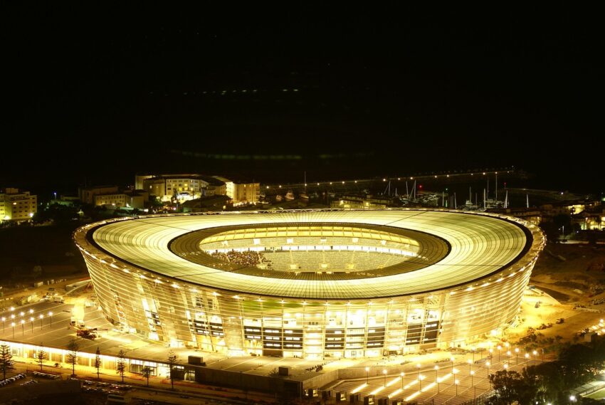 Stadium 1118445 1280 Triputi - Energieheilung für Mensch und Raum, Energetisches Feng Shui, Elementarkraft Metall, hell erleuchtetes Fußballstadion in Kapstadt, Dach erinnert an Frisbeering, Hauptfarben: gelb, schwarz, braun, grün